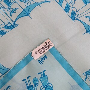 May include: A light blue silk scarf with a white border and a hand-drawn design of people in a city setting. The scarf has a label that reads "scarves by Ira, pure silk, designed + hand-screened in USA".