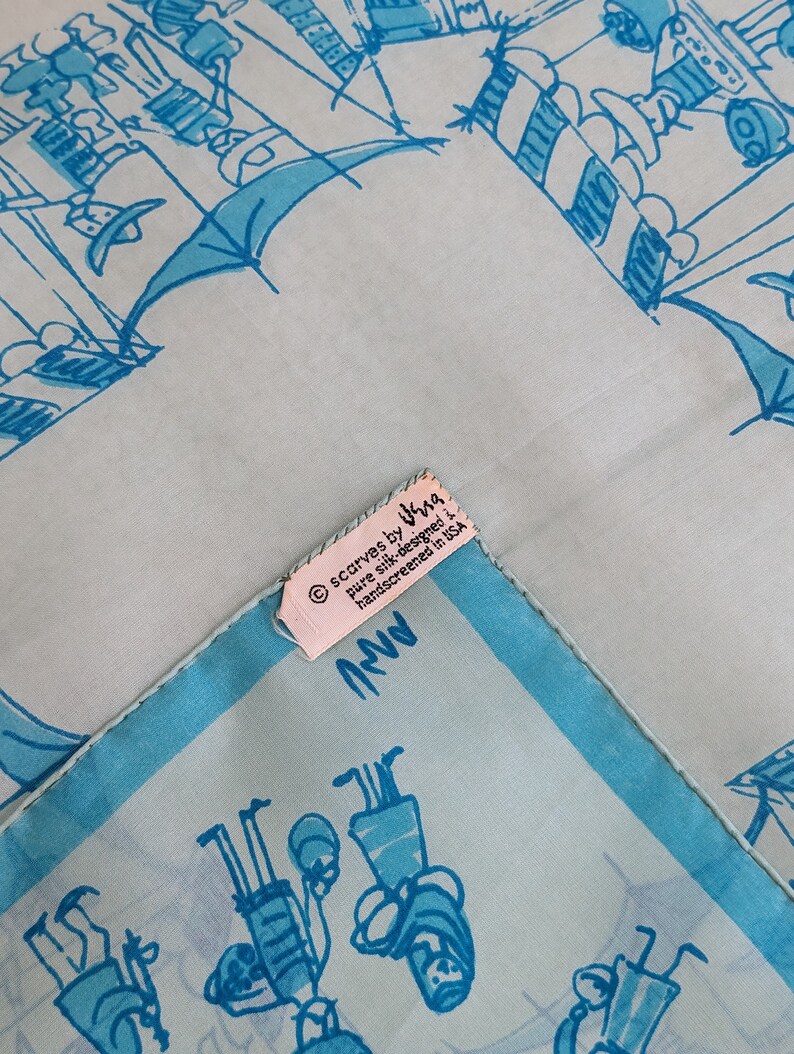 May include: A light blue silk scarf with a white border and a hand-drawn design of people in a city setting. The scarf has a label that reads "scarves by Ira, pure silk, designed + hand-screened in USA".