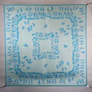 May include: A light blue square tablecloth with a repeating pattern of blue figures and geometric shapes. The figures are arranged in a border around the center of the tablecloth.