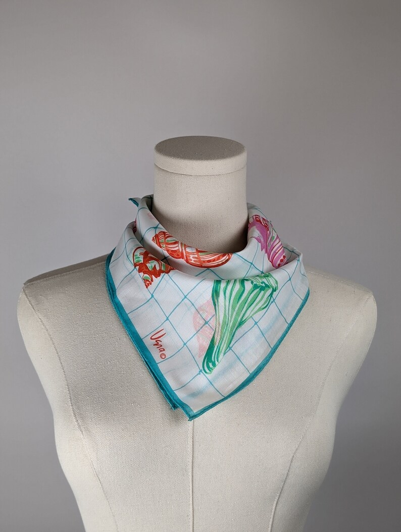 Op de afbeelding: Een witte katoenen bandana met een turquoise rand en een rasterpatroon. De bandana heeft een kleurrijke illustratie van groenten, waaronder een groene courgette, een rood-oranje paprika en een roze ui. De tekst "Vanna" is in rood op de bandana gedrukt.