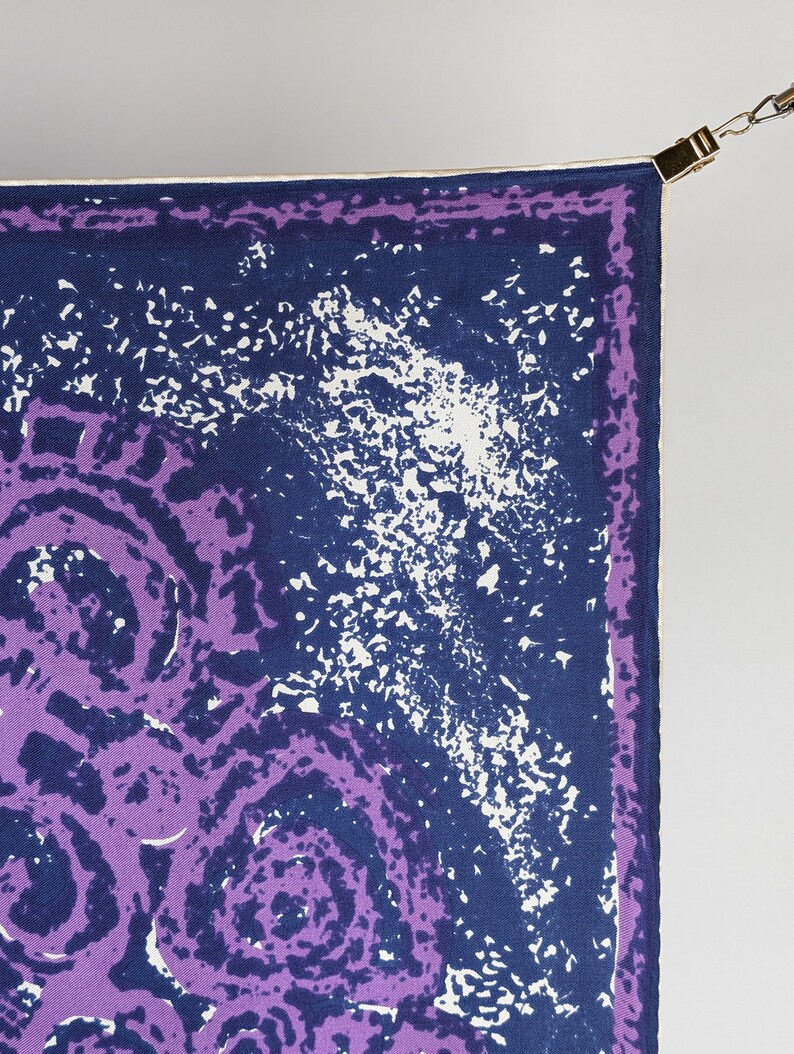 May include: A purple and blue patterned silk scarf with a white border. The scarf is hanging from a gold clip.