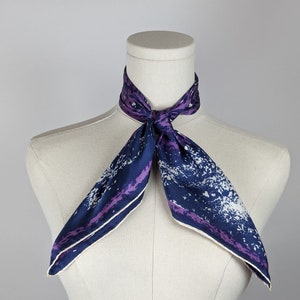 May include: A purple and blue silk scarf with a white and purple abstract print. The scarf is tied around a white mannequin's neck.