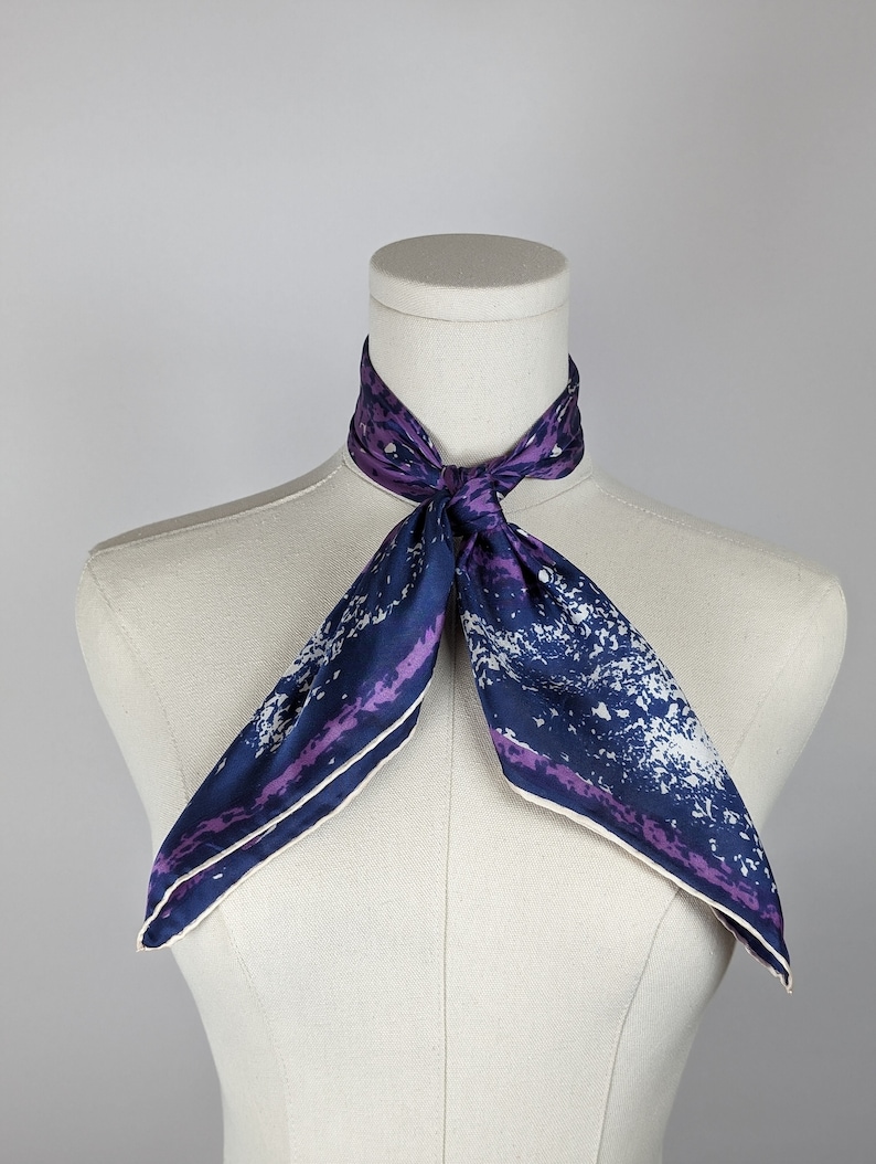 May include: A purple and blue silk scarf with a white and purple abstract print. The scarf is tied around a white mannequin's neck.