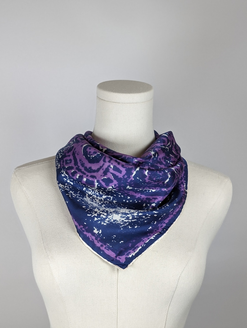 May include: A purple and blue patterned silk scarf with a white background. The scarf is folded into a triangle shape and is displayed on a white mannequin.
