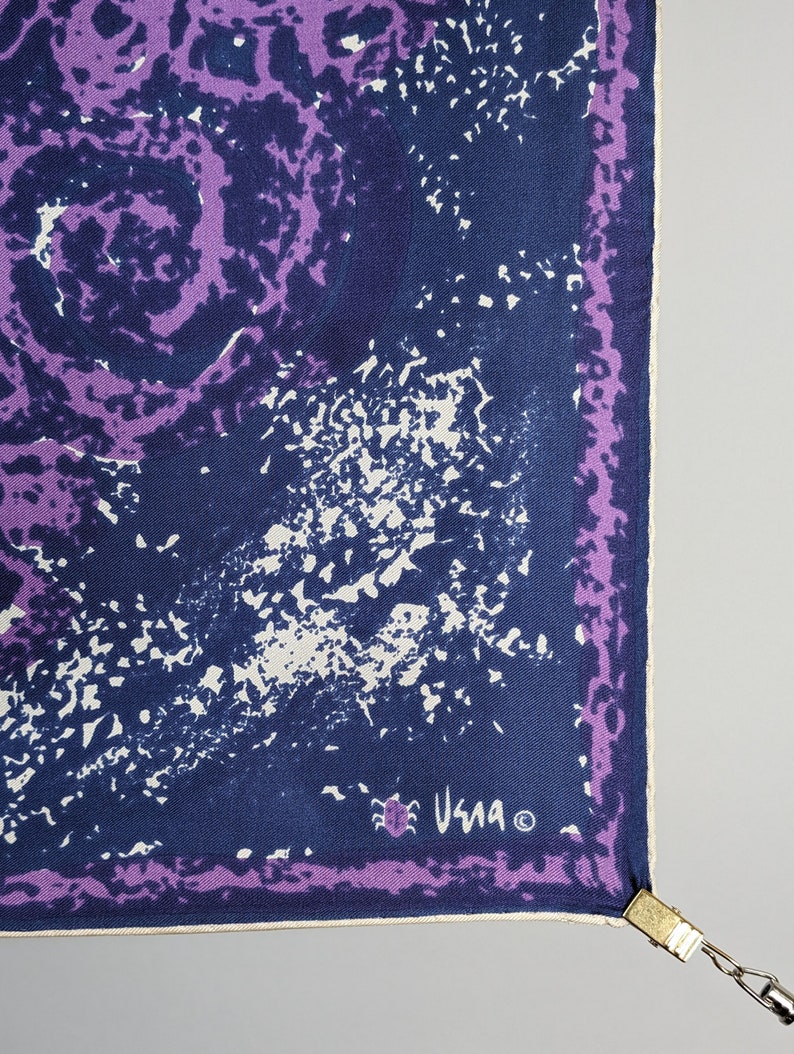 May include: A purple and blue silk scarf with a white and blue abstract design. The scarf has a white border and the signature "Usma" in the bottom right corner.