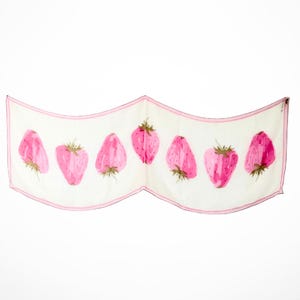 May include: A rectangular white scarf featuring a repeating pattern of pink strawberries with green stems. The scarf is bordered by a thin pink line along its edges and is displayed in a draped, wavy shape against a plain white background.