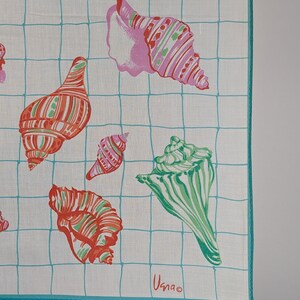 Op de afbeelding: Een wit katoenen zakdoek met een blauw rasterpatroon en kleurrijke schelpillustraties. Het zakdoek heeft een turquoise rand en de tekst "Usnao" is gedrukt in de rechterbenedenhoek.