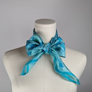 May include: A light blue silk scarf with a blue and white pattern of people and buildings. The scarf is tied around a white mannequin's neck.
