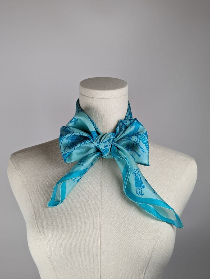 May include: A light blue silk scarf with a blue and white pattern of people and buildings. The scarf is tied around a white mannequin's neck.