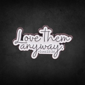May include: White and black sign with the words "Love them anyway" in a cursive font. The text "Luke 23:34" is below the main text. The sign is on a dark gray background.