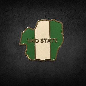 May include: A gold-edged pin in the shape of Imo State, Nigeria, with the state's flag colours: green, white, and green. The text "IMO STATE" is centered on the white stripe.