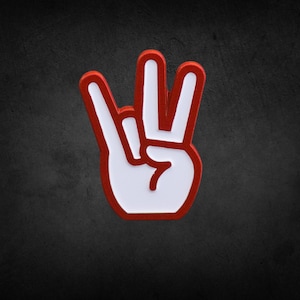May include: A white hand gesture pin with a red outline, displaying the peace sign. The pin is set against a dark, textured background. The design is bold and graphic, suitable for accessories.