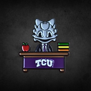 Puede incluir: Un pin de rana gris de dibujos animados con traje y corbata, detrás de un escritorio con una manzana roja y libros. El escritorio es morado con "TCU" en letras blancas. Fondo gris oscuro.