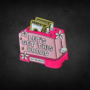 May include: A pink and red enamel pin shaped like a toaster with the text "LET'S GET THIS BREAD". The toaster has white flower and star accents, and two dollar bills are sticking out of the top. The bottom of the toaster says "HOT & FRESH".