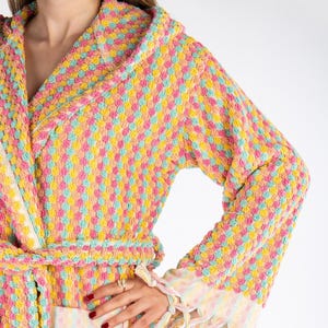 May include: A colourful hooded robe with a textured, dotted pattern in shades of yellow, pink, and blue. The robe has a matching belt and fringed trim on the sleeves and pockets. The robe is made of a soft, absorbent material.