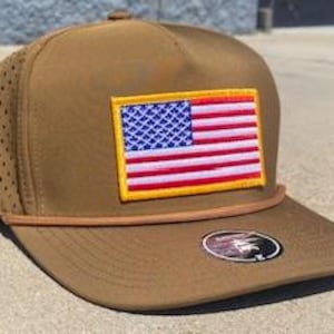 May include: A brown baseball cap with a flat peak and a mesh side panel. The cap features a rectangular patch with an American flag design and a brown rope detail. A small, circular emblem is on the peak.