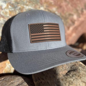May include: A gray and black trucker hat with a brown leather American flag patch. The hat has a black mesh back and a gray front.