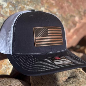May include: A navy blue and white trucker hat with a brown leather American flag patch. The hat has a mesh back and a Richardson 112 label.
