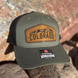 May include: A green and black trucker hat with a brown patch that says "COLORADO" and features a mountain range design. The hat is adjustable and has a Richardson 112 logo.