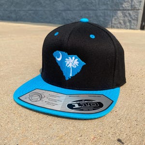 May include: Black and blue baseball cap featuring the South Carolina state flag. The cap has a flat brim and a Flexfit Tech label. The state flag is a blue palmetto tree and crescent on a blue outline.