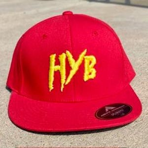 May include: A red baseball cap with a curved brim. The cap features the yellow embroidered letters "HYB" on the front. A small black and gold tag is on the brim.