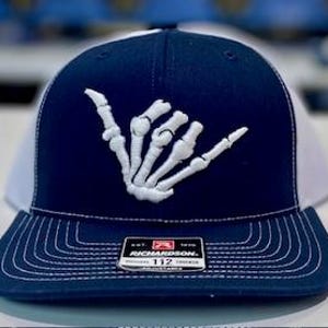 May include: Navy blue and white trucker hat with a white embroidered skeleton hand gesture. The hat features a structured front panel, a curved brim, and a mesh back. The Richardson brand patch is visible on the front.