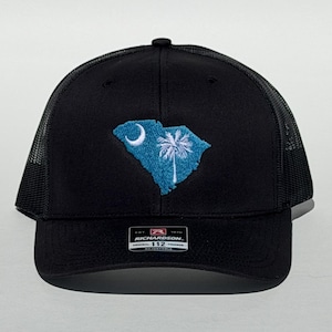May include: Black trucker hat with a mesh back and a curved brim. The front features an embroidered South Carolina state flag design in blue and white. The hat has an adjustable strap.