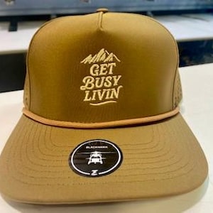 May include: A tan trucker hat with the text "GET BUSY LIVIN" in white above a mountain graphic. The hat has a flat brim, a brown rope detail, and a black and white circular logo on the brim.