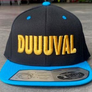 May include: Black and blue baseball cap with the word "DUUUVAL" embroidered in gold. The cap has a flat brim and a snapback closure. The cap has a blue brim and blue accents on the top.