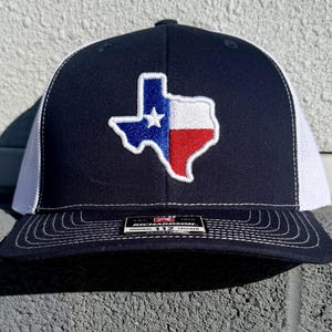 May include: Navy blue and white trucker hat featuring an embroidered Texas state flag. The Texas flag is in the center of the hat, with a white star, blue, white, and red colors. The hat has a curved brim and a mesh back.