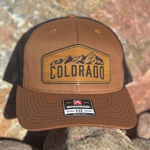 May include: Brown and black trucker hat with a brown leather patch that says "COLORADO" with a mountain range graphic. The hat is adjustable and has a black mesh back.