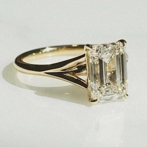 Könnte beinhalten: Ein goldener Verlobungsring mit einem Diamanten im Smaragdschliff. Der Ring hat ein geteiltes Schaftdesign. Der rechteckige Diamant ist klar und funkelnd. Der Ring ist vor einem weißen Hintergrund platziert.