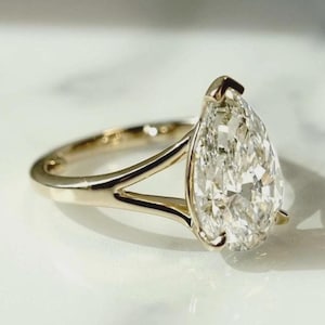 Könnte beinhalten: Ein goldener Ring mit einem großen, birnenförmigen Diamanten. Der Diamant ist klar und funkelnd. Der Ring hat ein geteiltes Schaftdesign.