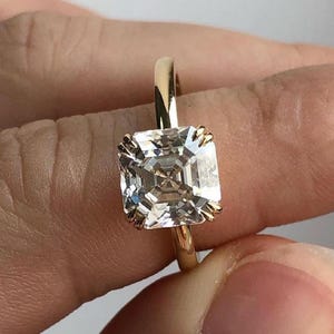 Puede incluir: Un anillo de oro con un gran diamante de talla cuadrada. El diamante transparente es multifacético y refleja la luz. El anillo tiene una banda de oro pulido. El diamante está sujeto por cuatro garras.