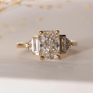 Könnte beinhalten: Ein goldener Ring mit einem großen, rechteckigen, klaren Edelstein als Mittelstück. Zwei kleinere, rechteckige Edelsteine flankieren den Hauptstein. Kleine, runde Edelsteine sind auf beiden Seiten des Rings gesetzt.