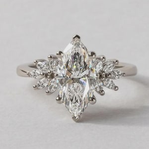 May include: A diamond engagement ring with a marquise-cut central stone and a halo of smaller diamonds. The ring has a silver band and a large, elongated diamond, encircled by a cluster of smaller, petal-shaped diamonds. The design is elegant and striking.