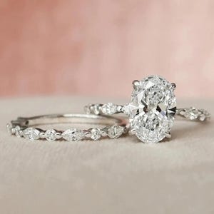 May include: Three silver rings with sparkling white gemstones. The main ring features a large oval-cut stone, while the other two have smaller, round and marquise-cut stones. The rings are set against a soft pink background.