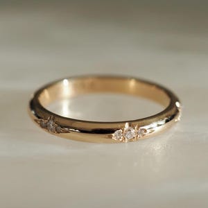 May include: A gold ring featuring small gemstones. The ring has a smooth, polished band and is decorated with three clusters of tiny, clear stones. The ring is set against a soft, neutral background, highlighting its elegant design.