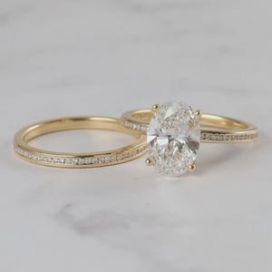 May include: A pair of gold rings. One ring has a large oval-cut diamond, the other is a plain band. Both rings have small diamonds set into the bands. The rings are displayed on a white surface.