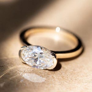 May include: A gold ring featuring a large, oval-cut diamond. The diamond is set in a bezel setting, and the band is smooth and polished. The ring is displayed on a neutral surface, with soft lighting.