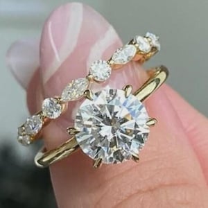 May include: Two gold rings with sparkling white gemstones. One ring features a large, round-cut stone, while the other has smaller, pear-shaped stones. The rings are set against a soft pink background.
