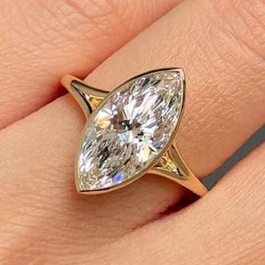 May include: A gold ring featuring a marquise-cut diamond. The diamond is set in a bezel setting, and the band has a split design. The ring is displayed on a finger, against a neutral background.