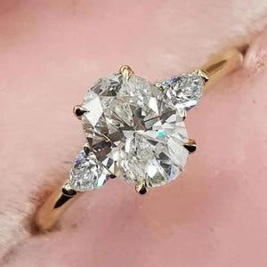 May include: A gold engagement ring featuring a large oval-shaped diamond and two smaller pear-shaped diamonds on the side.