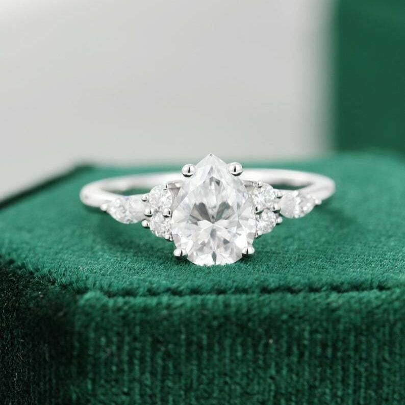 May include: A silver ring with a pear-shaped, clear gemstone as the focal point. Smaller, round gemstones flank the main stone on either side. The ring is set against a green velvet surface.