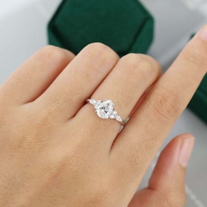 May include: A silver ring with a pear-shaped, clear gemstone as the centerpiece, flanked by smaller, round gemstones. The ring is set on a hand, with a dark green velvet box in the background.