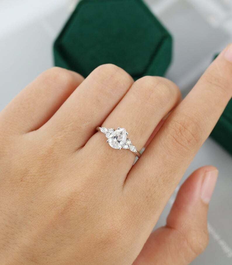 May include: A silver ring with a pear-shaped, clear gemstone as the centerpiece, flanked by smaller, round gemstones. The ring is set on a hand, with a dark green velvet box in the background.
