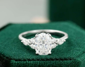 Peut inclure: Une bague de fiançailles en argent avec une pierre centrale de forme ovale et des pierres plus petites sur la bande. La bague est posée sur un écrin en velours vert.