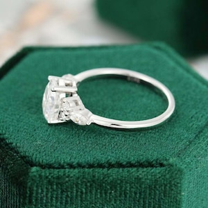 May include: A silver engagement ring with a large, oval-cut diamond as the centerpiece. The ring features smaller, accent diamonds on either side of the main stone. The ring is set on a green velvet surface.