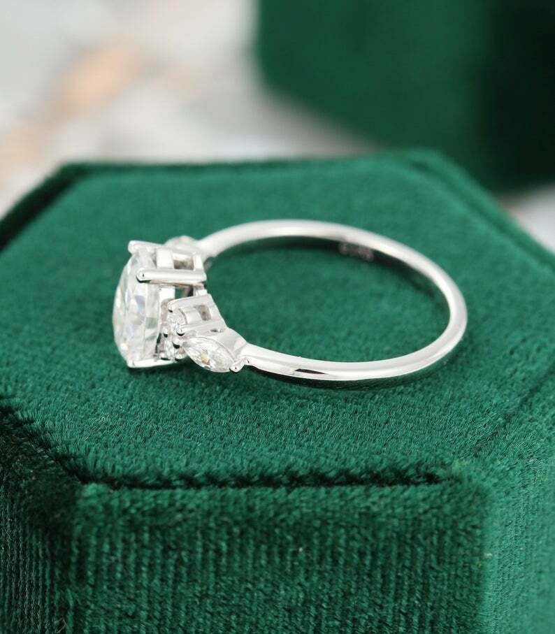 May include: A silver engagement ring with a large, oval-cut diamond as the centerpiece. The ring features smaller, accent diamonds on either side of the main stone. The ring is set on a green velvet surface.