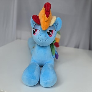 Puede incluir: Un juguete de peluche azul claro con una melena y cola de arcoíris multicolor. El personaje tiene ojos grandes y expresivos con pestañas largas, una pequeña sonrisa y orejas puntiagudas. El cuerpo de tela suave es de color azul sólido.